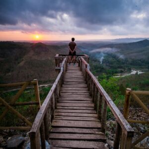 A person reflecting on a wooden boardwalk at sunset, overlooking a vast mountainous landscape.