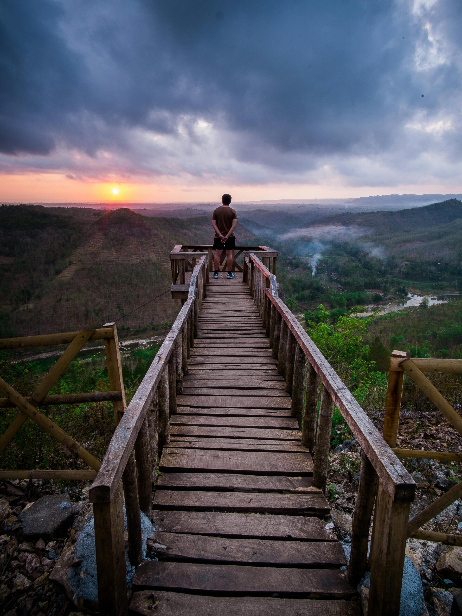 A person reflecting on a wooden boardwalk at sunset, overlooking a vast mountainous landscape.